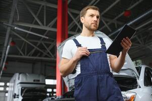 Bearded mechanic in overalls standing in garage of a car salon and holding tablet. He is about to diagnostic breakdown. photo