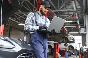 A bearded auto-mechanic in working uniform is standing next to a broken car and using the special laptop for car maintenance. An auto-mechanic in workshop repairs car. photo
