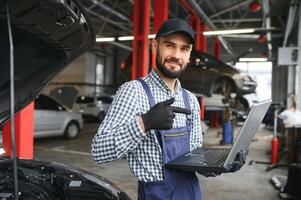 A bearded auto-mechanic in working uniform is standing next to a broken car and using the special laptop for car maintenance. An auto-mechanic in workshop repairs car. photo