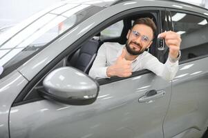 Handsome young businessman in classic white suit is smiling, looking at camera and showing car keys while sitting in car in a motor show. photo