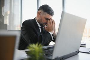 Tired stressed office worker sitting at desk and thinking, he is rubbing his eyes and feeling exhausted. photo