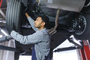 Indian car mechanic standing and working in service station. Car specialists examining the lifted car. Professional repairmen wearing mechanic uniform in blue color. photo