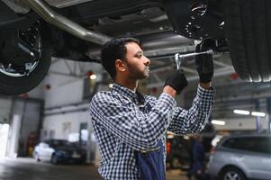 Indian car mechanic standing and working in service station. Car specialists examining the lifted car. Professional repairmen wearing mechanic uniform in blue color. photo