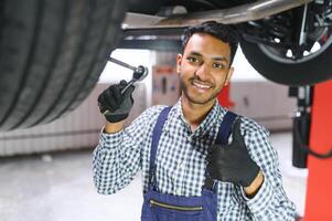 Indian car mechanic standing and working in service station. Car specialists examining the lifted car. Professional repairmen wearing mechanic uniform in blue color. photo