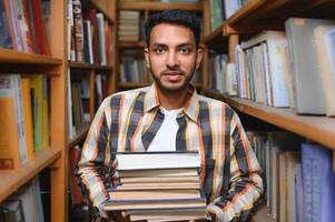 Portrait of cheerful male international Indian student with backpack, learning accessories standing near bookshelves at university library or book store during break between lessons. Education concept photo