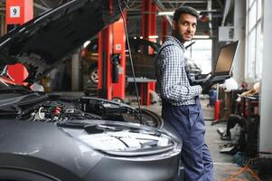 Mechanic man mechanic manager worker using a laptop computer checking car in workshop at auto car repair service center. Engineer young man looking at inspection vehicle details under car hood. photo