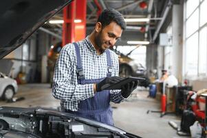 Mechanic man mechanic manager worker using a laptop computer checking car in workshop at auto car repair service center. Engineer young man looking at inspection vehicle details under car hood. photo