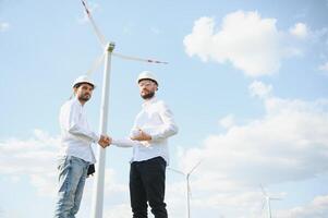 Close up of indian technician and inspector shaking hands while standing on farm with wind turbines. Concept of people, teamwork and eco friendly energy photo