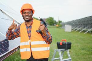 Competent energy engineer in grey overalls and orange helmet checking solar panels while walking on field. African american man carrying clipboard and container with instruments photo