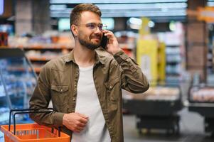 Quick text during shopping. Handsome young man holding mobile phone and smiling while standing in a food store photo