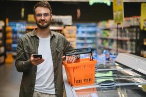 Quick text during shopping. Handsome young man holding mobile phone and smiling while standing in a food store photo