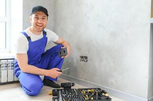Smiling professional electrician in uniform replacing a socket in apartment after renovation work photo