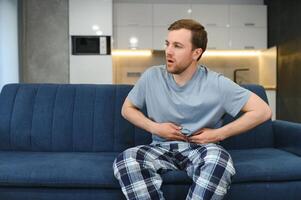 Guy with a beard feeling unwell with a stomach ache while sitting in a couch at home photo