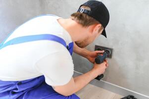 Confident professional electrician in uniform using screwdriver while replacing a socket in apartment after renovation work photo