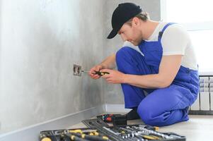Smiling professional electrician in uniform replacing a socket in apartment after renovation work photo