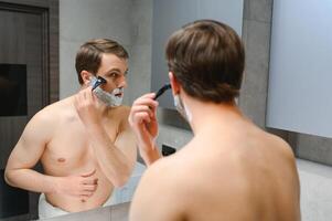 Handsome young man is shaving his face and looking at the mirror. photo