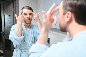 Handsome man looking at mirror and applying moisturizing cream on cheeks in bathroom. Groomed young guy doing skincare morning routine after taking a shower. photo