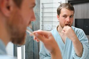 Brushing teeth in the morning. Rear view of handsome young beard man brushing his teeth and smiling while standing against a mirror. photo