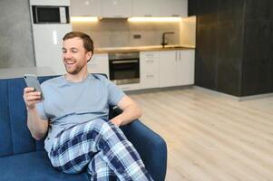 A happy smiling caucasian guy, in casual stylish clothes, sits on the couch at home, holds a phone and a banking card in his hands, makes online payment for purchases, enters a card number photo