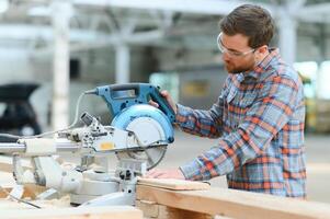 Professional carpenter using sawing machine for cutting wooden board at sawmill. Skilled cabinet maker working with electric circular saw at woodworking workshop photo