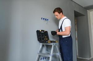 Electrician Builder at work, installation of sockets and switches. Professional in overalls with an electrician's tool. Against the background of the repair site. photo