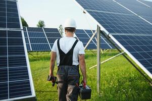 masculino trabajador en uniforme con un perforar - destornillador en su mano en un escalera de tijera cerca solar paneles foto