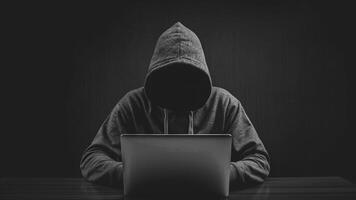 anonymous hacker in hoodie with no face using laptop computer on dark background. thief get into internet system. cyber crime and digital fraud prevention photo