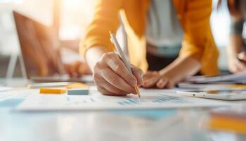 Person writing on a paper at a desk with charts and documents. A collaborative office work environment with sunlight streaming in. photo