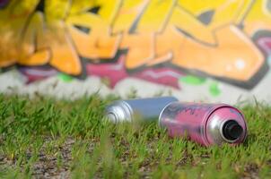 Several used spray cans with paint and caps for spraying paint under pressure on grass near the painted wall in colored graffiti photo