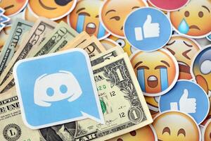 KYIV, UKRAINE - APRIL 20, 2024 Discord logo and many usd money bills. Symbol of famous social network platform and dollar currency banknotes on lot of emoji reactions photo