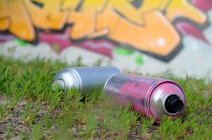 Several used spray cans with paint and caps for spraying paint under pressure on grass near the painted wall in colored graffiti photo
