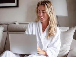 Smiling woman in a white bathrobe using a laptop while seated on a comfortable couch in a cozy living room setting. photo