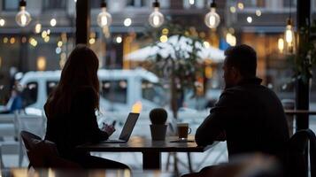 Silhouettes of two people sitting at a cafe table with laptops, surrounded by hanging lights in a cozy urban setting. photo