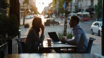 Two people having a conversation at an outdoor cafe table with a laptop and coffee as the sun sets on a bustling street. photo