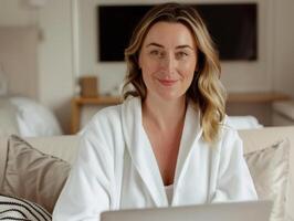 Smiling woman in a white bathrobe using a laptop while seated on a comfortable couch in a cozy living room setting. photo
