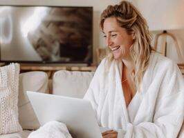 Smiling woman in a white bathrobe using a laptop while seated on a comfortable couch in a cozy living room setting. photo