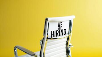 White office chair with We Are Hiring text displayed on its backrest, set against a vibrant yellow background, symbolizing job opportunities and recruitment. photo