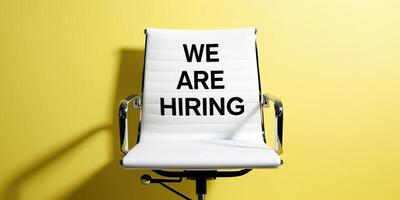 White office chair with We Are Hiring text displayed on its backrest, set against a vibrant yellow background, symbolizing job opportunities and recruitment. photo