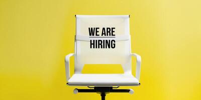 White office chair with We Are Hiring text displayed on its backrest, set against a vibrant yellow background, symbolizing job opportunities and recruitment. photo