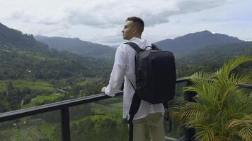 Man with view of tropical mountain valley. Action. Man on observation deck in tropical mountains. Man with backpack on sightseeing attraction overlooking green mountains photo