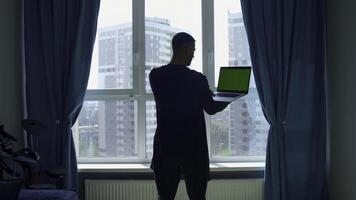 Rear view of a male student studying at home and using his laptop. Media. Man standing by the window and looking at green chroma key screen. photo