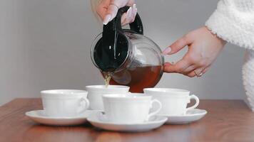 Close-up of a person pouring tea from a glass kettle into white cups on a wooden table. The moment of serving tea, highlighting the simplicity and elegance of the setting. video
