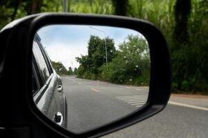 View of mirrors wing. Rear view of a gray car with asphalt road. At the end is a curved path with a symbol. Beside with green forest. speed bumps on the road. Under view of blue sky. photo