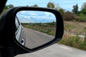 Inside view of mirrors wing. Rear view of a gray car with asphalt road. And open space on the side of the road. Revealing distant communities in the daytime. Under blue sky at day time. photo