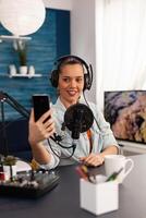 Portrait of blogger woman taking selfie for audience using smartphone working in podcast home studio. Content creator recording new fashion and beauty review and having fun on social media platform. photo