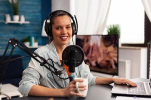 Smiling influencer woman sitting in front of camera recording for fashion blog. Digital blogger vlogger streaming talk show in studio using headphones, professional podcast microphone photo