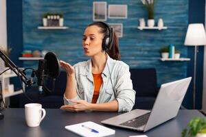 Vlogger giving flying kiss while making daily online blog. Digital influcencer recording talk show at home studio brodcast using headphones, professional podcast microphone and modern laptop photo