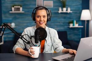 Portrait of smiling blogger looking at camera before starting live at home studio podcast holding cup of coffee. Content creator blogger woman recording brodcast live streaming for Internet photo