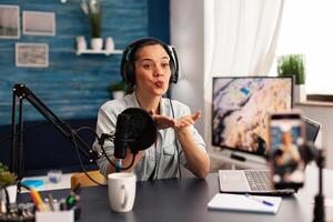 Influencer giving flying kiss while making new fashion series. Creative vlogger recording blog concept speaking and looking at smartphone on tripod home studio podcast photo