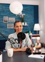 Smiling social media star sitting in front of camera and filming for youtube channel. Vlogger speaking and recording online talk show at home studio using modern equipment for digital podcast. photo